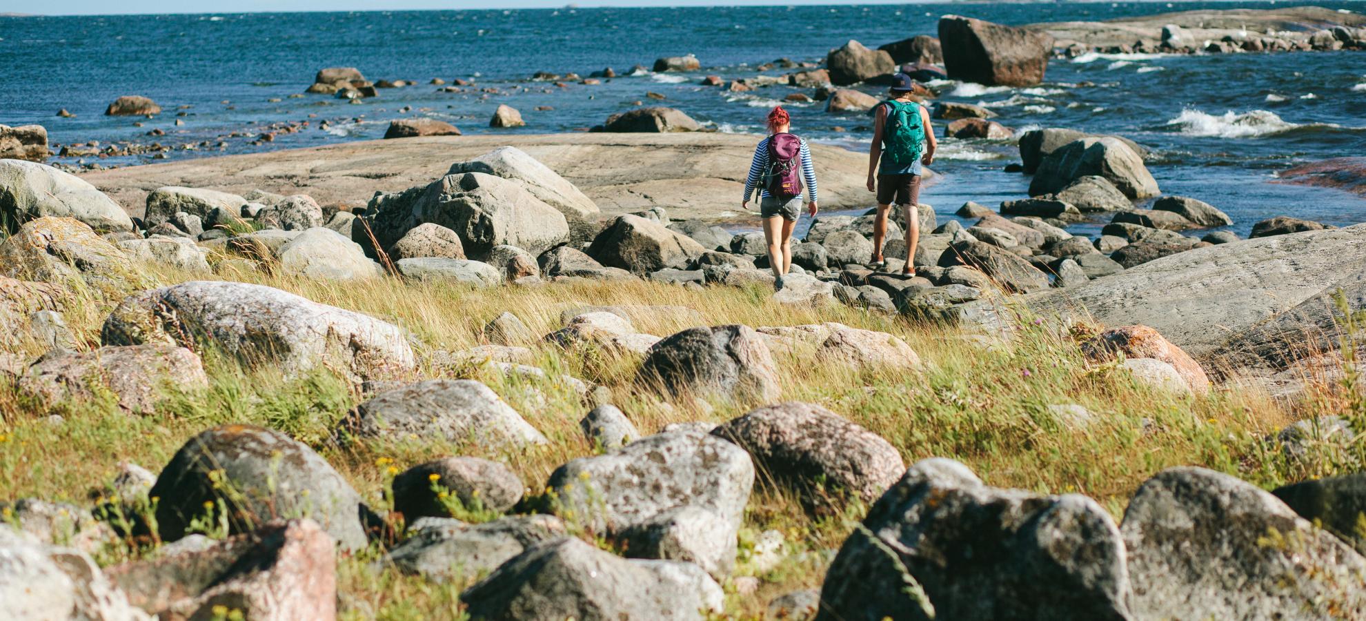 Two people are walking away from the camera along the grassy and rocky shore at Kaunisaari, the sea opening up in front of them towards the horizon. 
