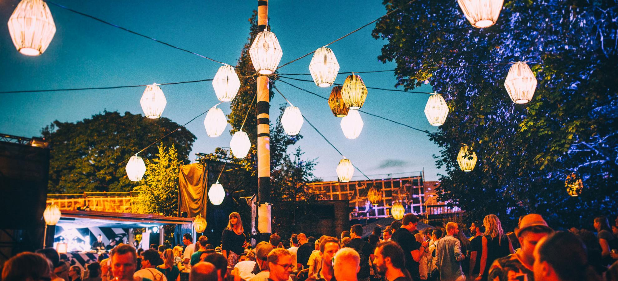 Large crowds gather during the evening at Flow Festival underneath dozens of paper lamps. A few large trees surround the area under a deep blue night's sky. 