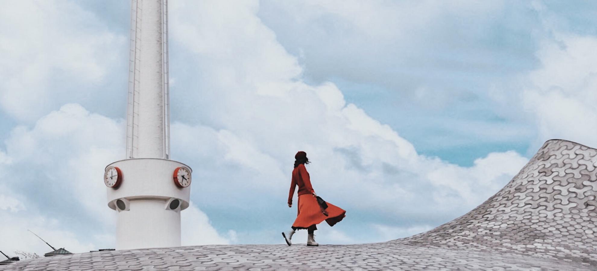Silvia Rodríguez Bernal walking across the top of one of Amos Rex's skylight domes, the tower of Lasipalatsi standing to the left in front of a mostly cloudy sky.