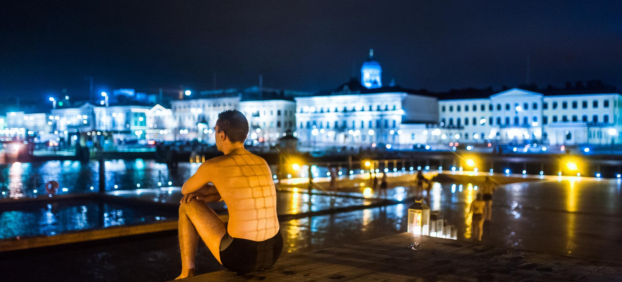 Sitting on the deck of Allas Sea Pool at night, the Predential Palace and the building of Pohjoisesplanadi lit up in the background, a man in swimming shorts looks out towards the harbour on the left.