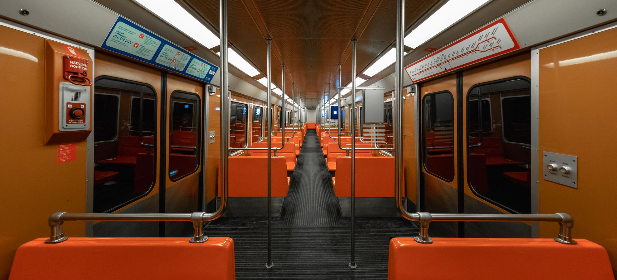 A view down the long carriage of an 80s metro train, bright orange bench seats line either side with the walls a more subdued shade of orange, florescent strip lights also running the length of the roof either side.