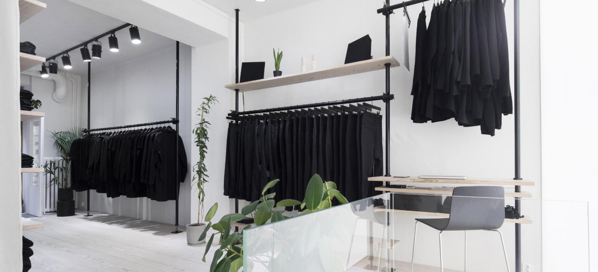 In a brilliant white store interior, a series of three black clothes on racks against the far wall are filled with various black clothing.