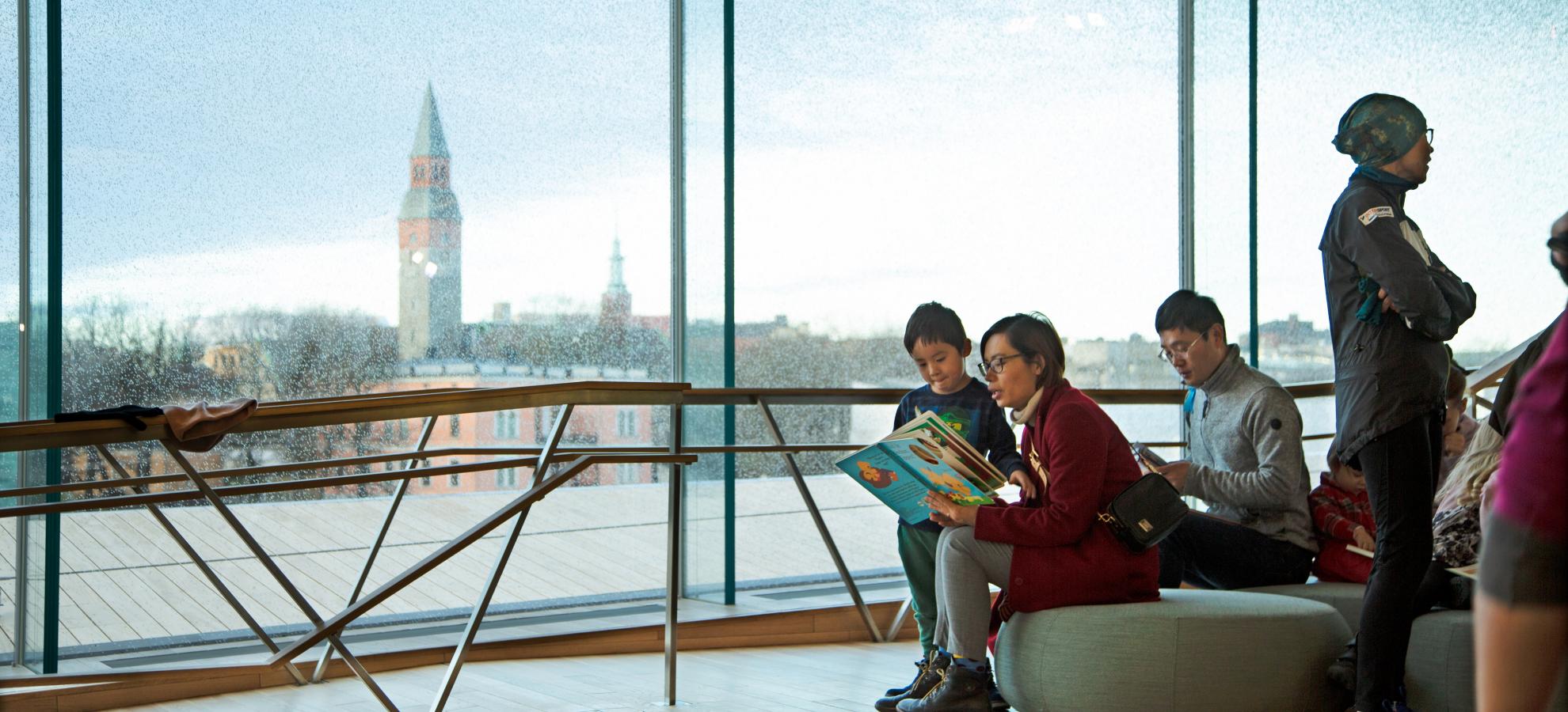 A woman is sitting and reading a book to a boy at Helsinki Central Library Oodi. Through the large windows behind them, can be seen the rooves of Töölö against a blue sky.