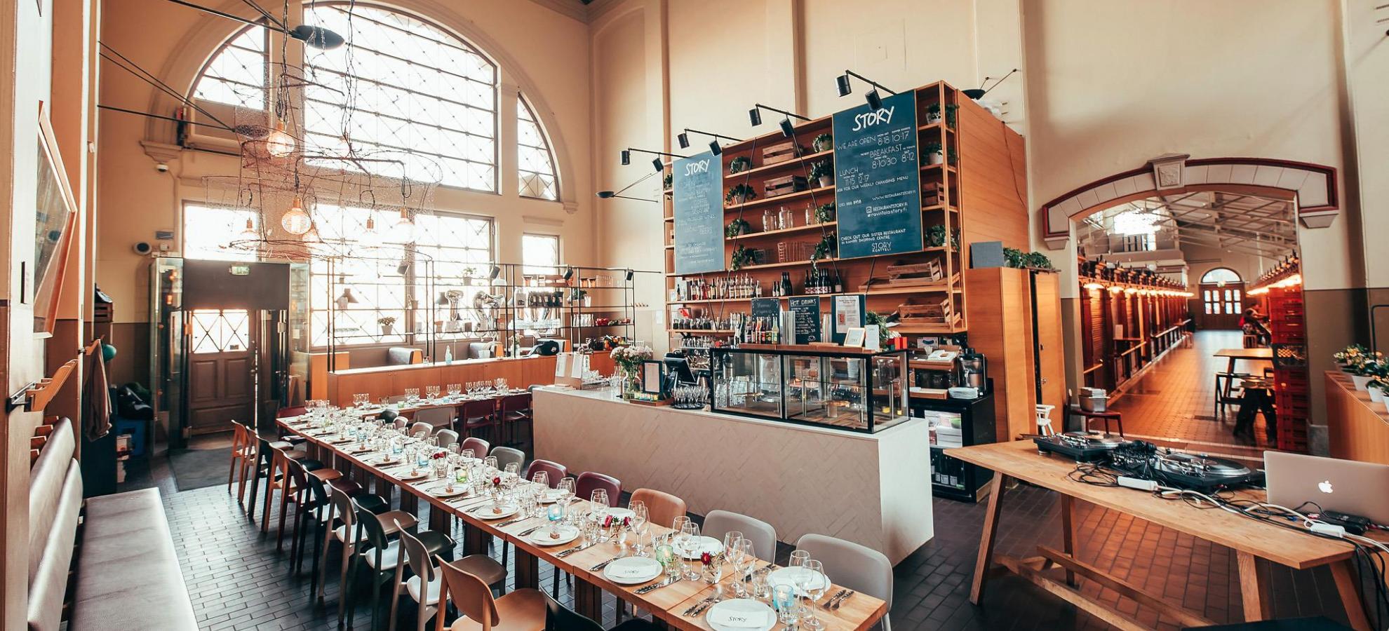 Story restaurant inside Old Market Hall, a tall, open space with large windows towards the left and the main market leading away down a corridor to the right of the restaurant's main counter. Behind the counter are a set of tall, wide shelves whereas in front of the counter, a long table has been set with dozens of chairs for a private event.