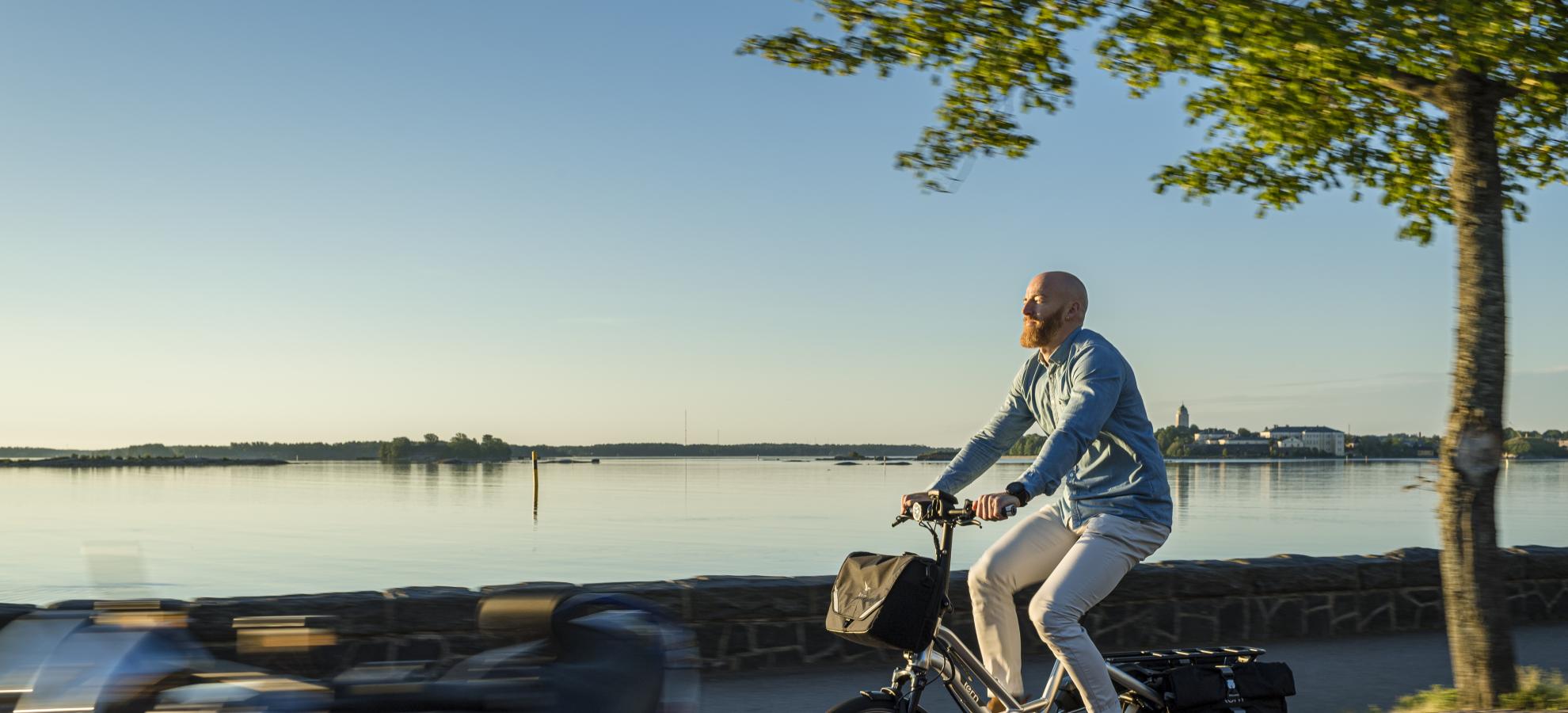 A man is cycling to the left of the photo on the path running by the Kaivopuisto shore, where the sea beyond is flat calm and the sun is shining.