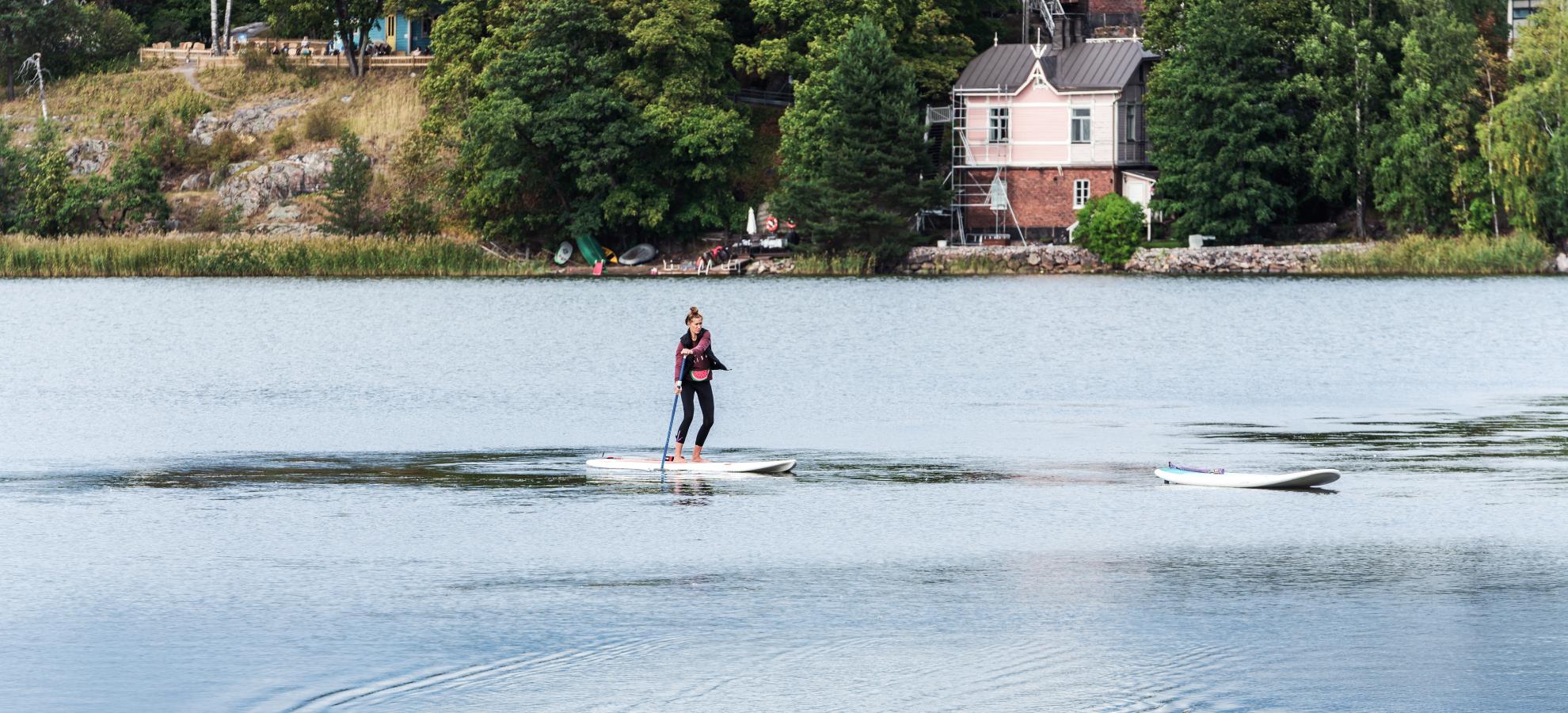 With three ducks swimming in the foreground, a woman paddles on a SUP board across Töölönlahti. The shoreline can be seen behind her, where a house stands surrounded by trees. 