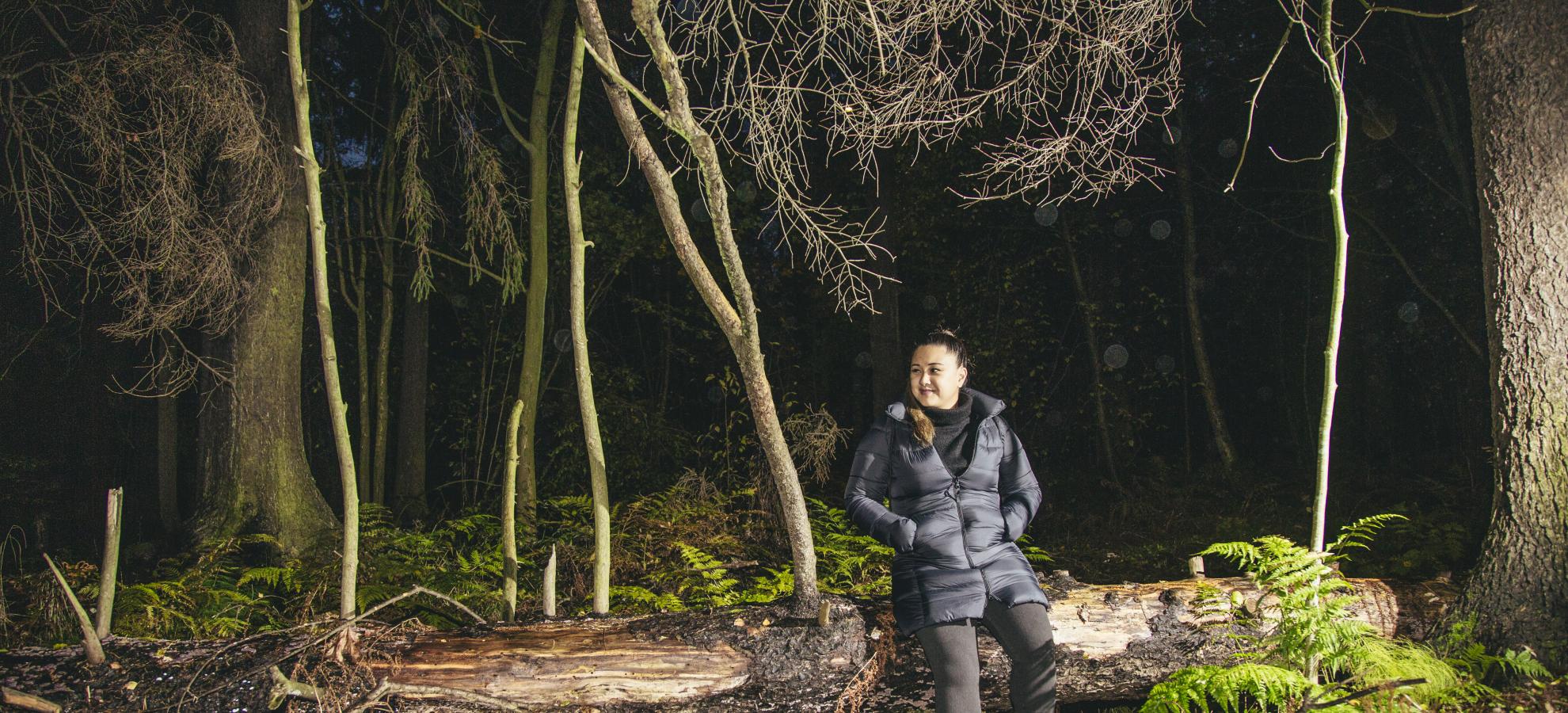 Adele Asuncion leaning against a fallen tree in the woods, looking to the left.