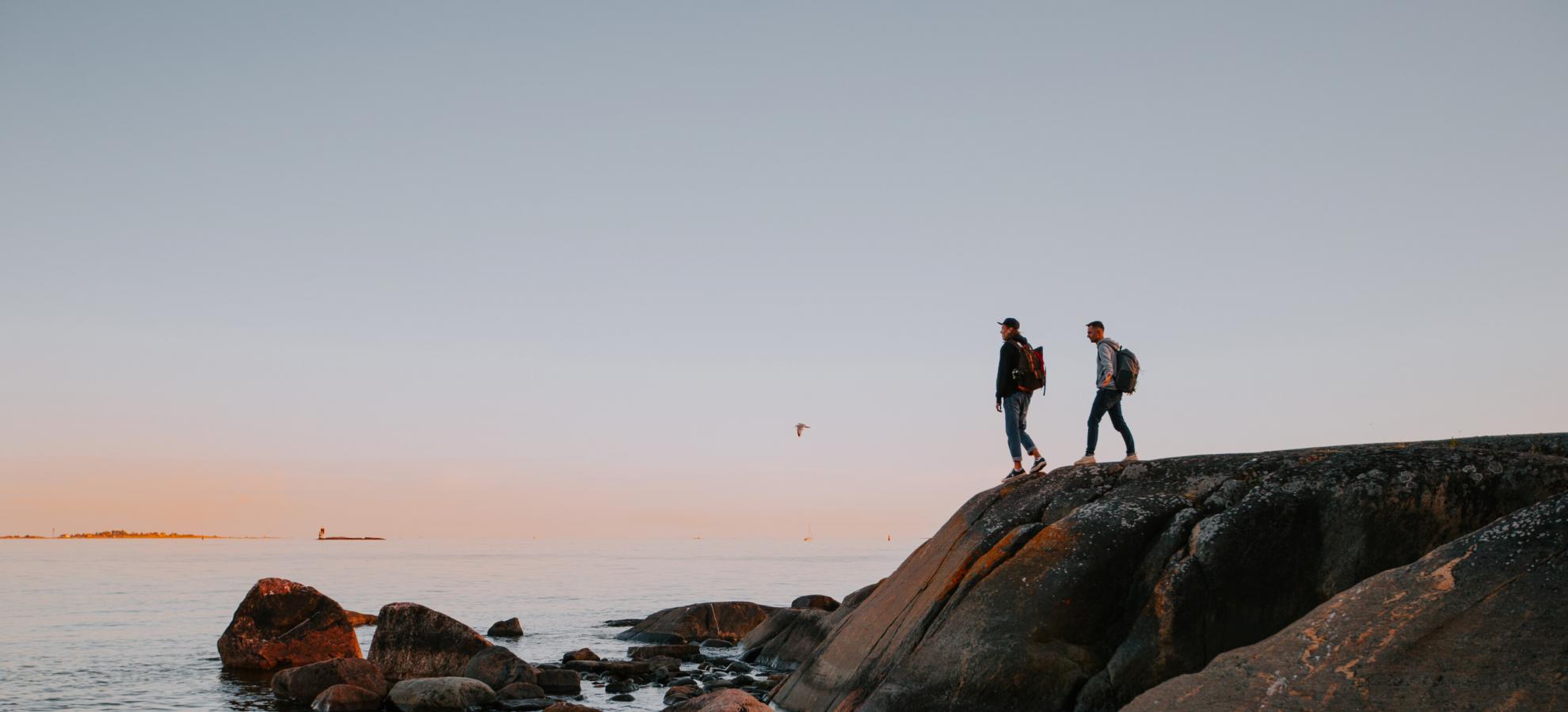 Towards the right of the picture, two men walk to the edge of some tall rocks on Pihlajsaari island and look out towards the sea at dusk.