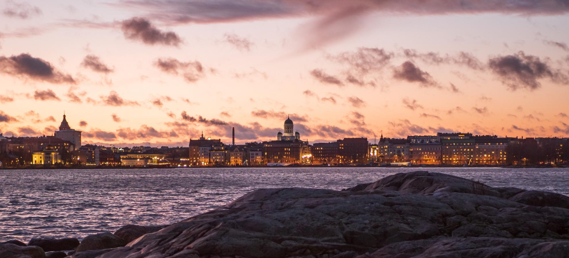 A view of Helsinki from a rocky shoreline during dusk with a pink sky. Helsinki Cathedral can be seen on top of the cityscape on the coastline in the distance. 