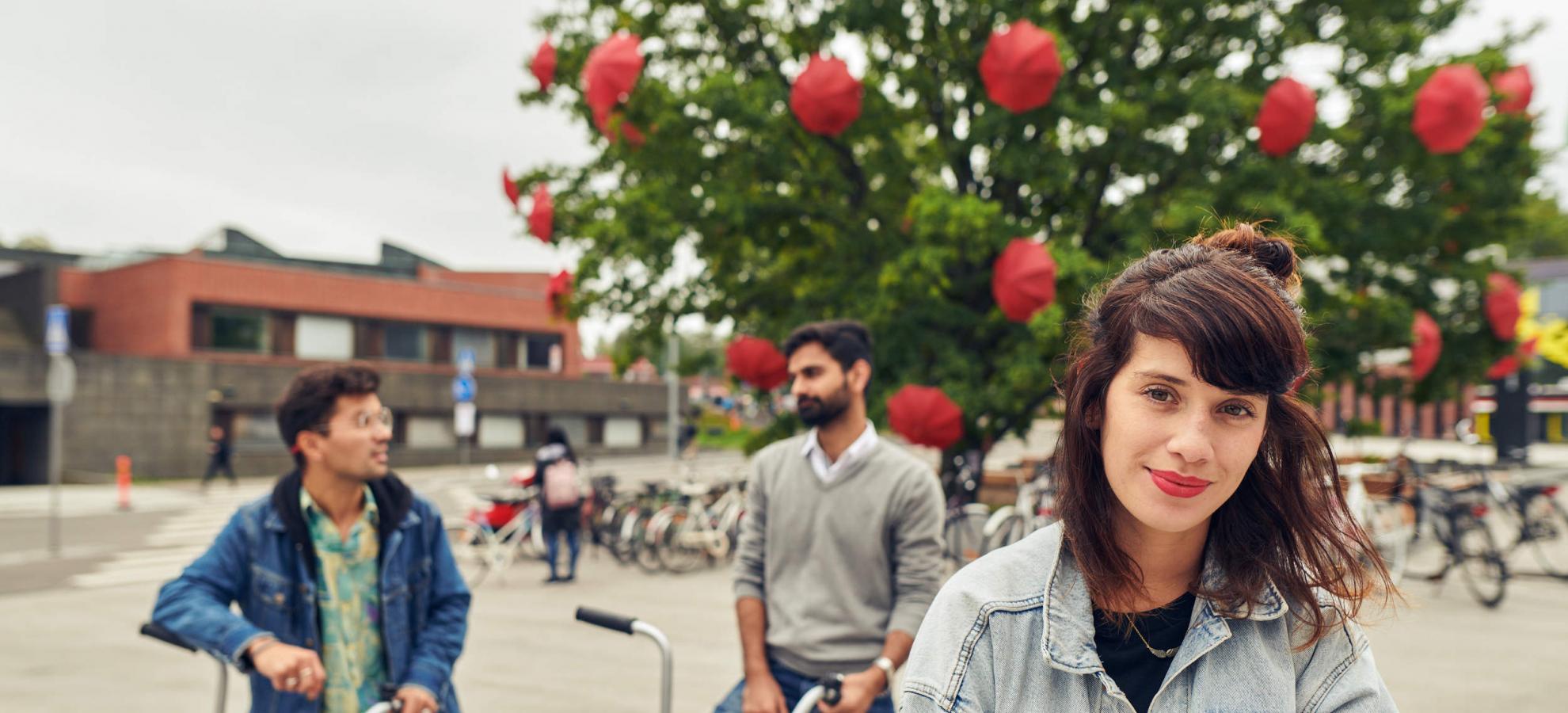 Three people are standing in front of Aalto University, a woman in the foreground looking straight at the camera, while 2 men mounted on bikes are chatting behind her. Behind them all are bike racks, a tree in full bloom dotted with red umbrellas, and the Aalto University buildings.