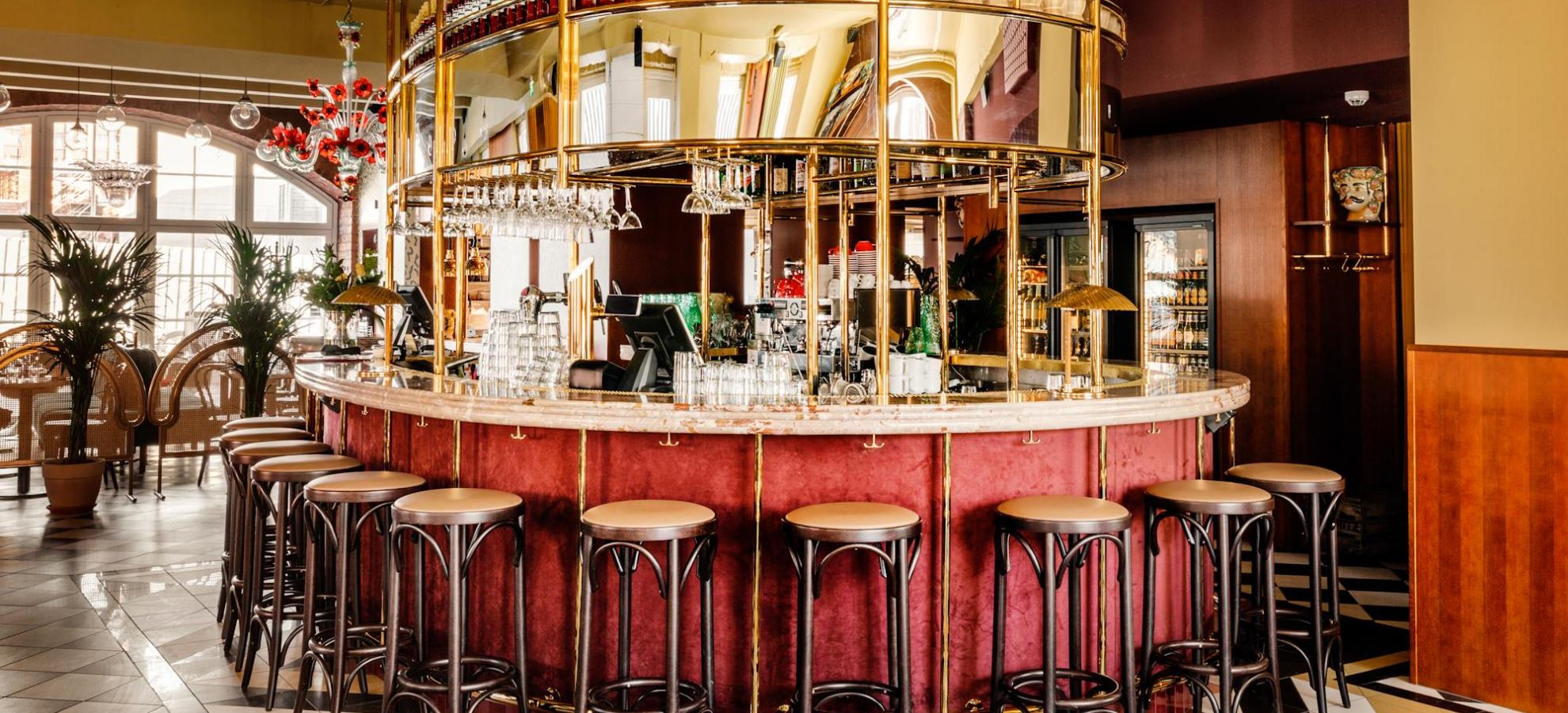 Ravintola Madonna bar, a fancy red walled bar topped with marble, bar stools surrounding it, stands in the center of a large room with a tiled floor.