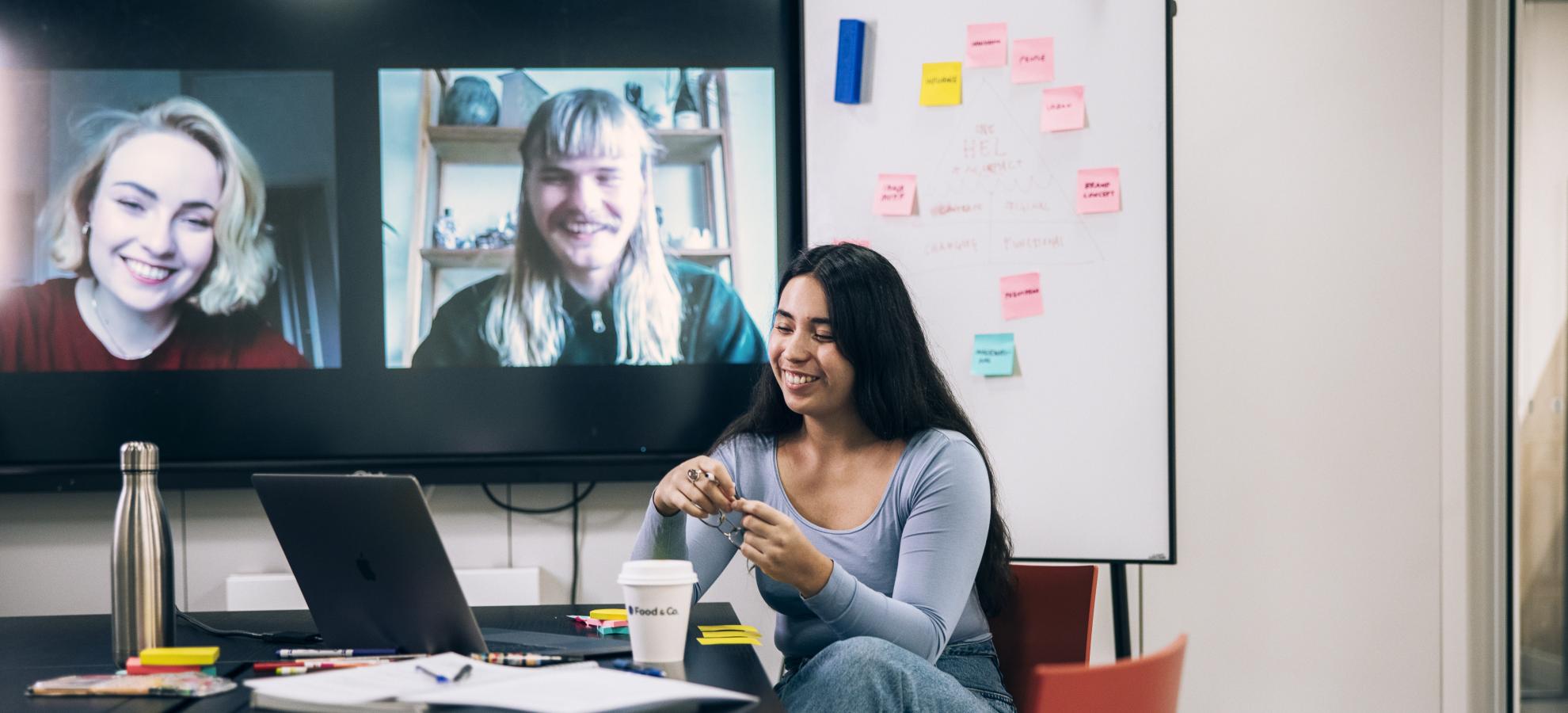 A woman sits at a large desk in a meeting space in Oodi library while having a virtual team meeting. Her teammates are displayed on a large TV screen in the background next to a whiteboard partially covered in sticky-notes.