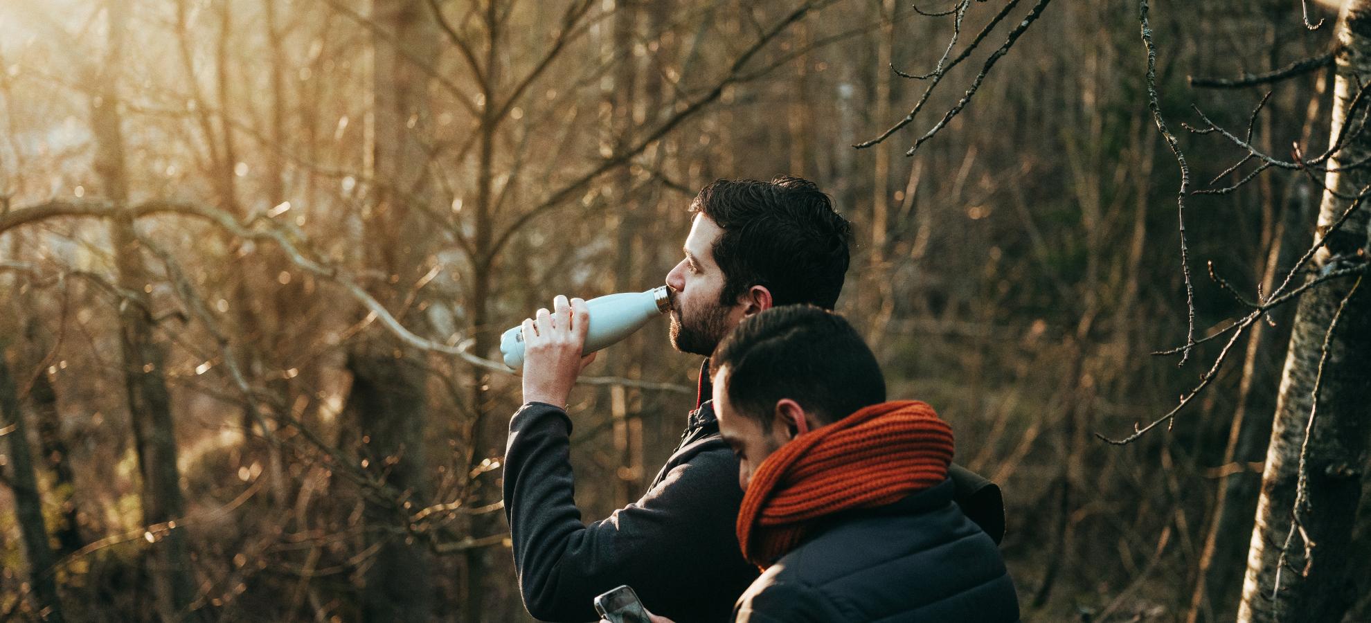 Two gentlemen, one drinking from a water bottle and the other checking their phone ,are standing in the woods with soft, golden sunlight shining through the trees towards them.