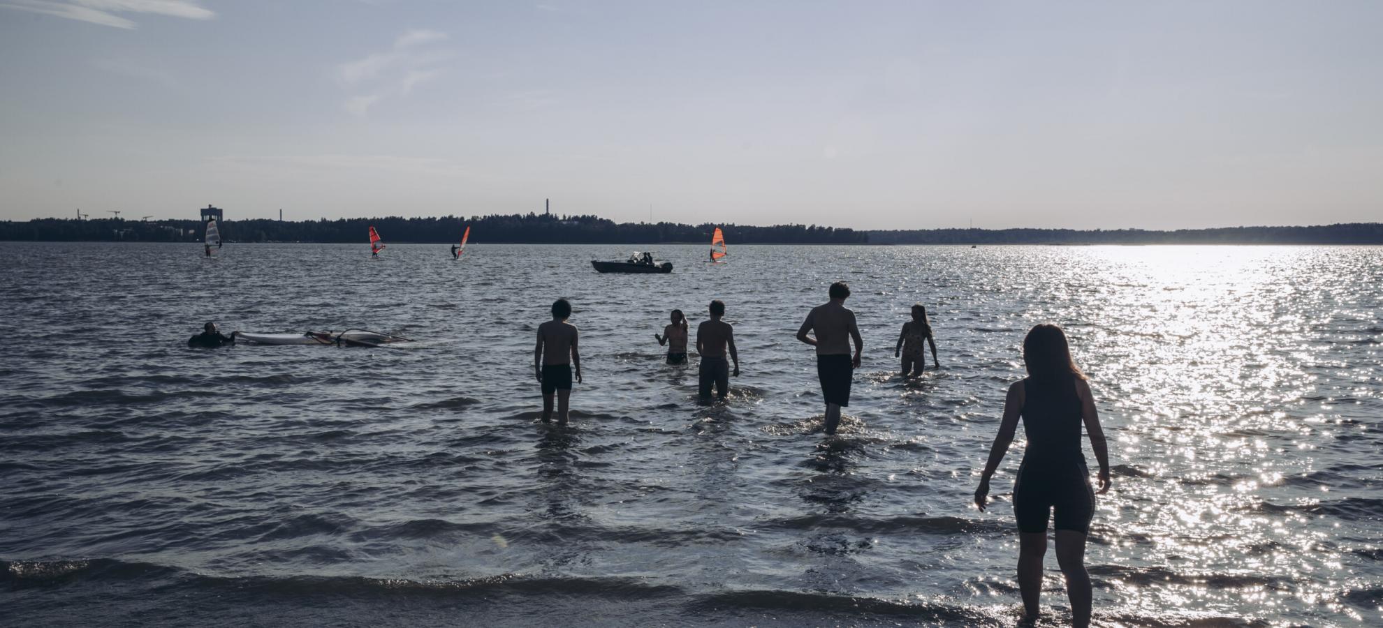 On a warm summer's day, sun glaring off the sea, a small group of people wade out into the water from the beach at Kasinonpuisto. The Servinniemi shoreline can be seen in the distance.