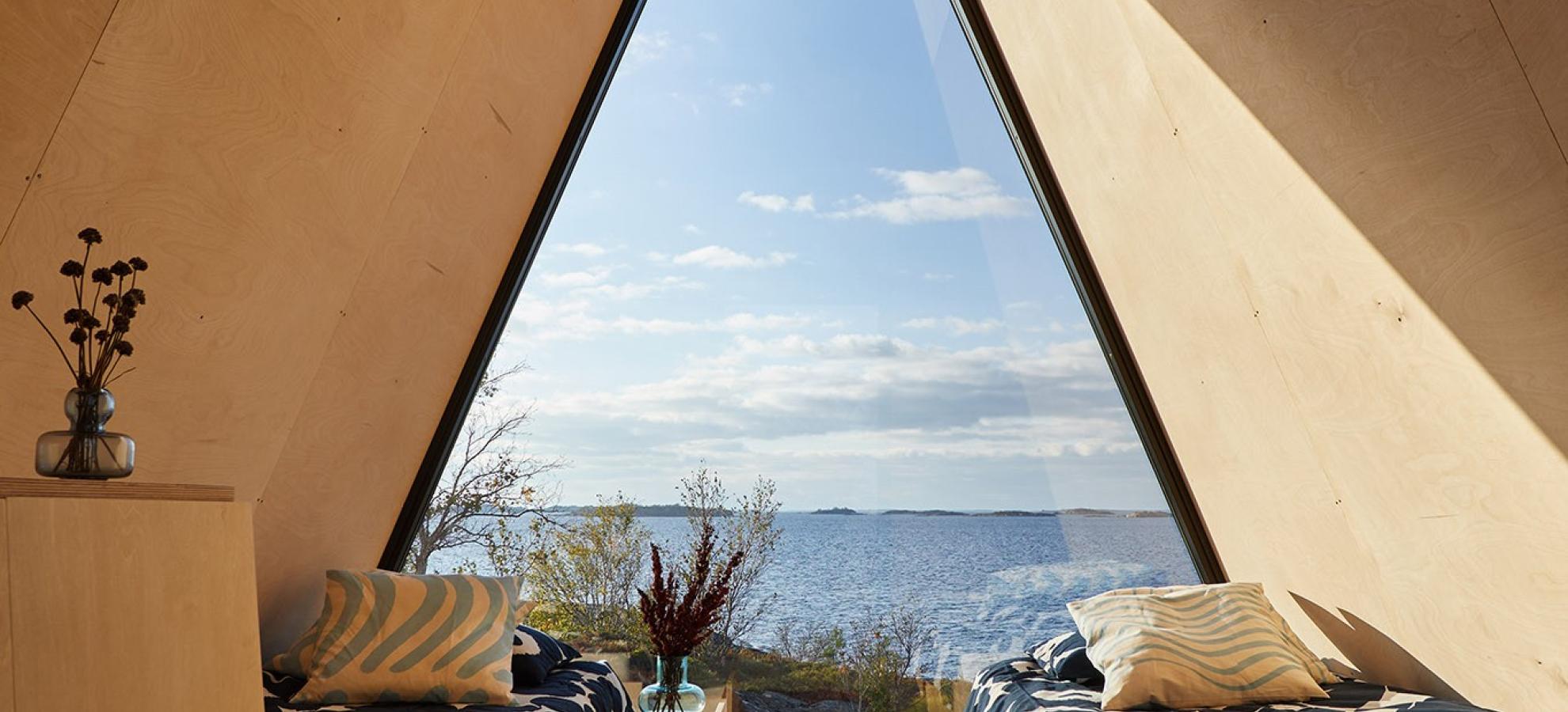 View looking out from a Nolla cabin, a wood constructed cabin that is prism shaped, with 2 single beds and a wall-sized triangular window at its end. The window overlooks a beautiful view of the sea