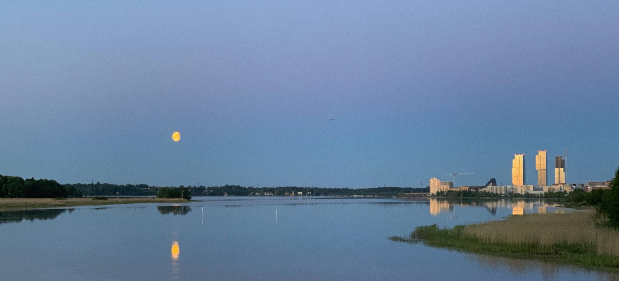 With a yellow moon against a deep blue sky at dusk, the bay next to Lammassaari stretches to shoreline on the horizon, where the tall buildings of Kalasatama standing on the right are lit by the setting sun.