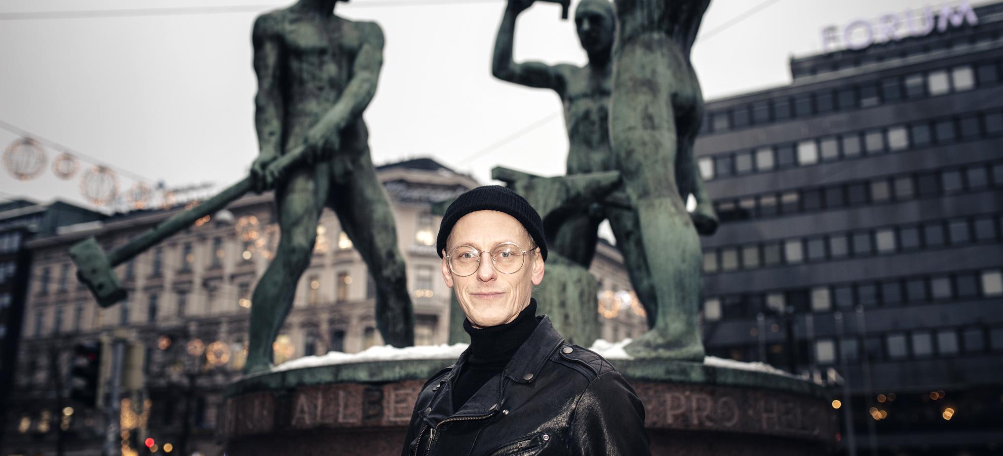 A man with eyeglasses and black beanie and leather jacket standing in front of a statue
